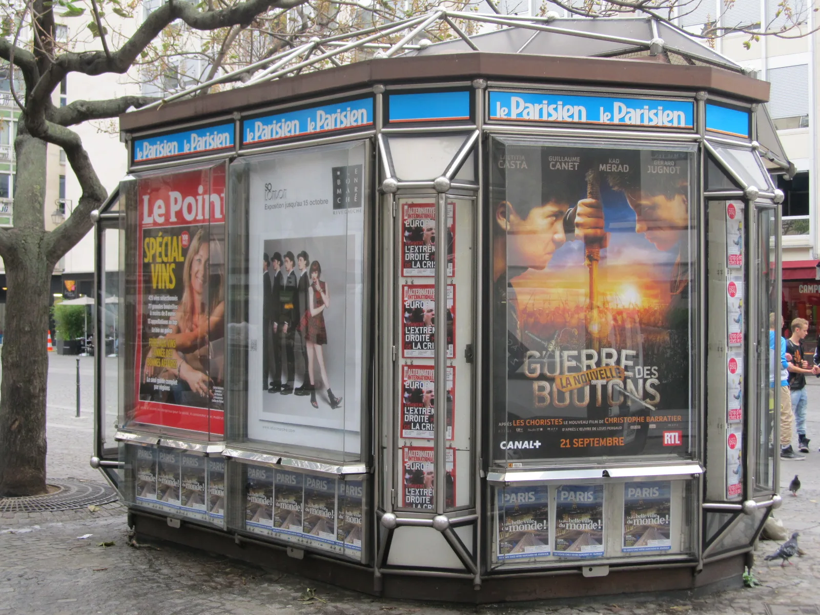 A Paris newsstand displays newspaper and magazine covers alongside posters.