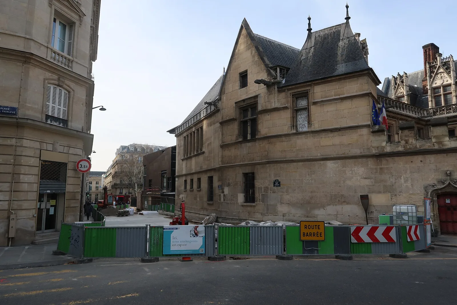 Construction barriers close a Paris street beside a stone building; a road-closed sign faces pedestrians.