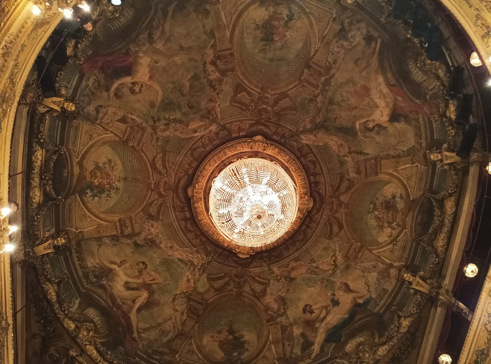 A chandelier hangs at the centre of an ornate painted theatre ceiling.