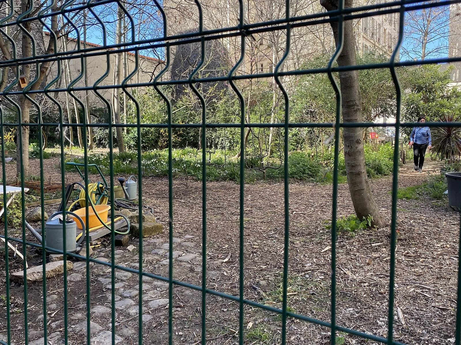 A metal fence borders the wooded Bois Dormoy community garden, with watering cans visible inside.